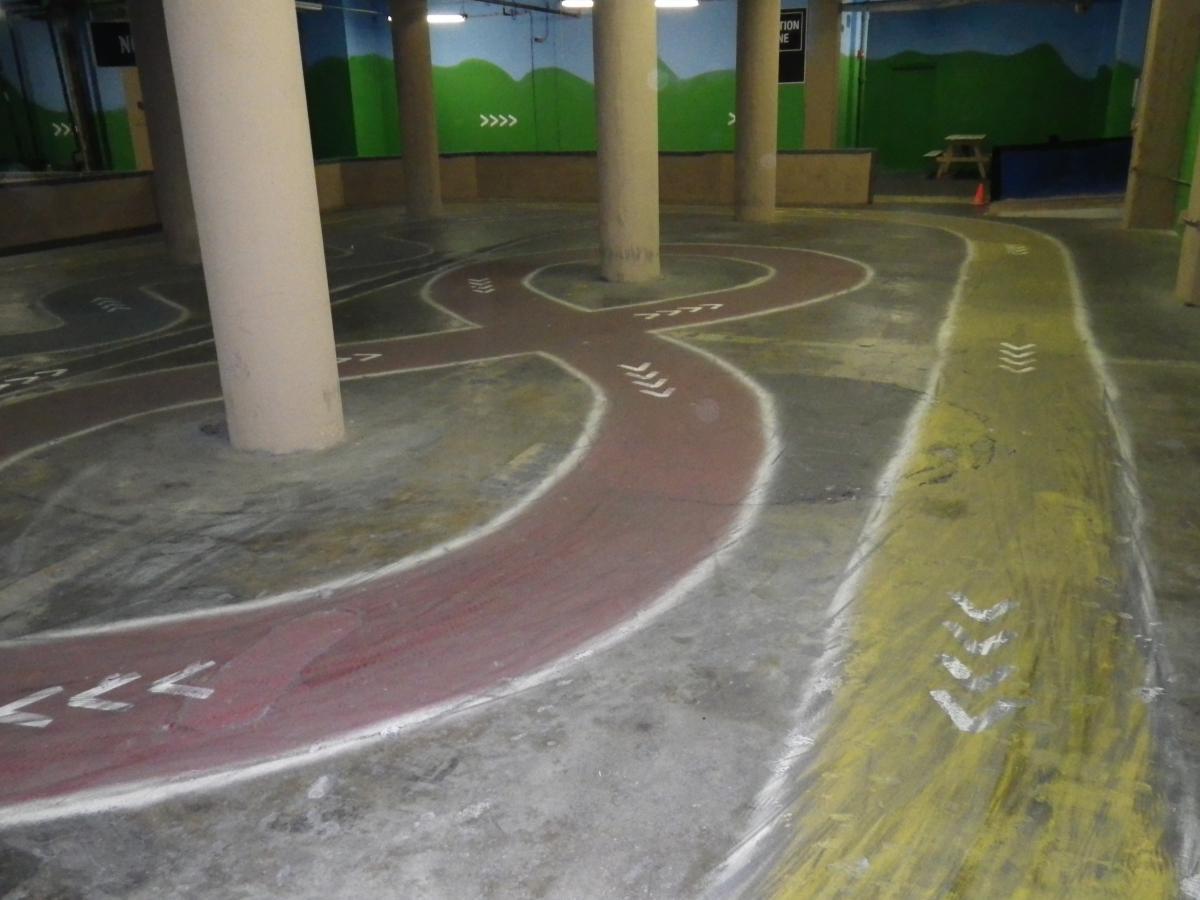 An indoor go-kart track with painted lanes in red and yellow, featuring white directional arrows. The space is surrounded by concrete pillars, with green-themed mural artwork on the walls in the background. Rays Indoor Mtb Park mountain bike trail.