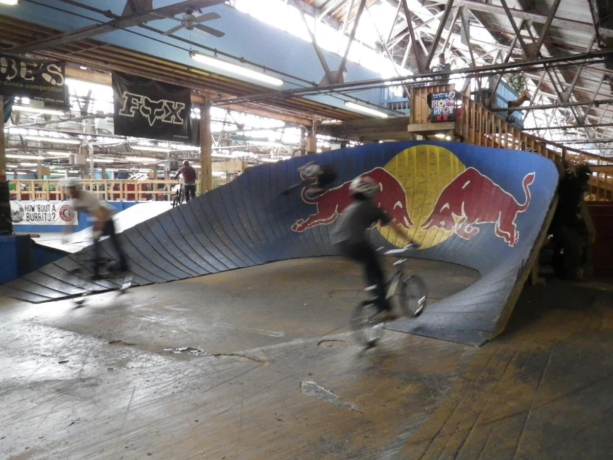 A dynamic indoor skate park scene featuring two riders on BMX bikes navigating a curved ramp decorated with the Red Bull logo. The setting includes a wooden structure in the background, with various banners hanging from the ceiling, creating an energetic and vibrant atmosphere. Rays Indoor Mtb Park mountain bike trail.