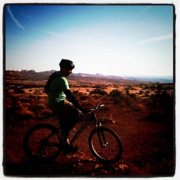 A mountain biker in a green shirt and helmet pauses on their bike, overlooking a desert landscape under a clear blue sky. The scene features rocky terrain and sparse vegetation in the background. Moab Brand Trails mountain bike trail.