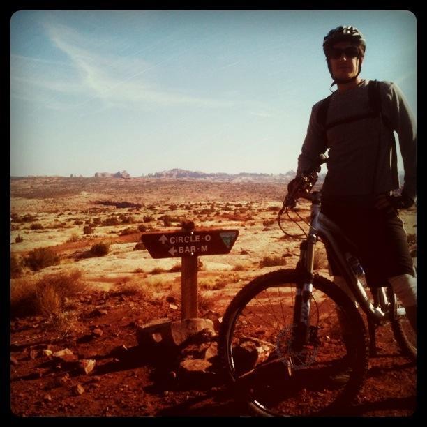 A mountain biker stands next to a trail sign pointing to "Circle-O" and "Bar-M," with a vast desert landscape and rocky formations in the background. The sky is clear, indicating sunny weather. Moab Brand Trails mountain bike trail.