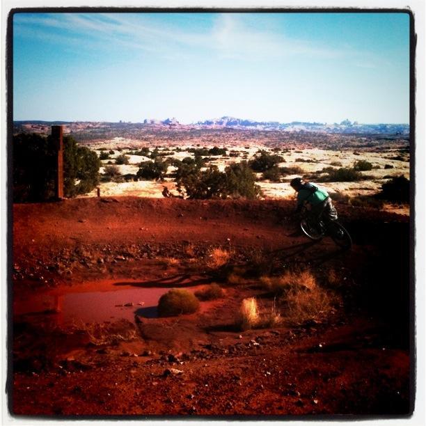 A mountain biker navigating a dirt track with a small puddle in the foreground, set against a backdrop of a vast, rocky landscape under a clear blue sky. Moab Brand Trails mountain bike trail.