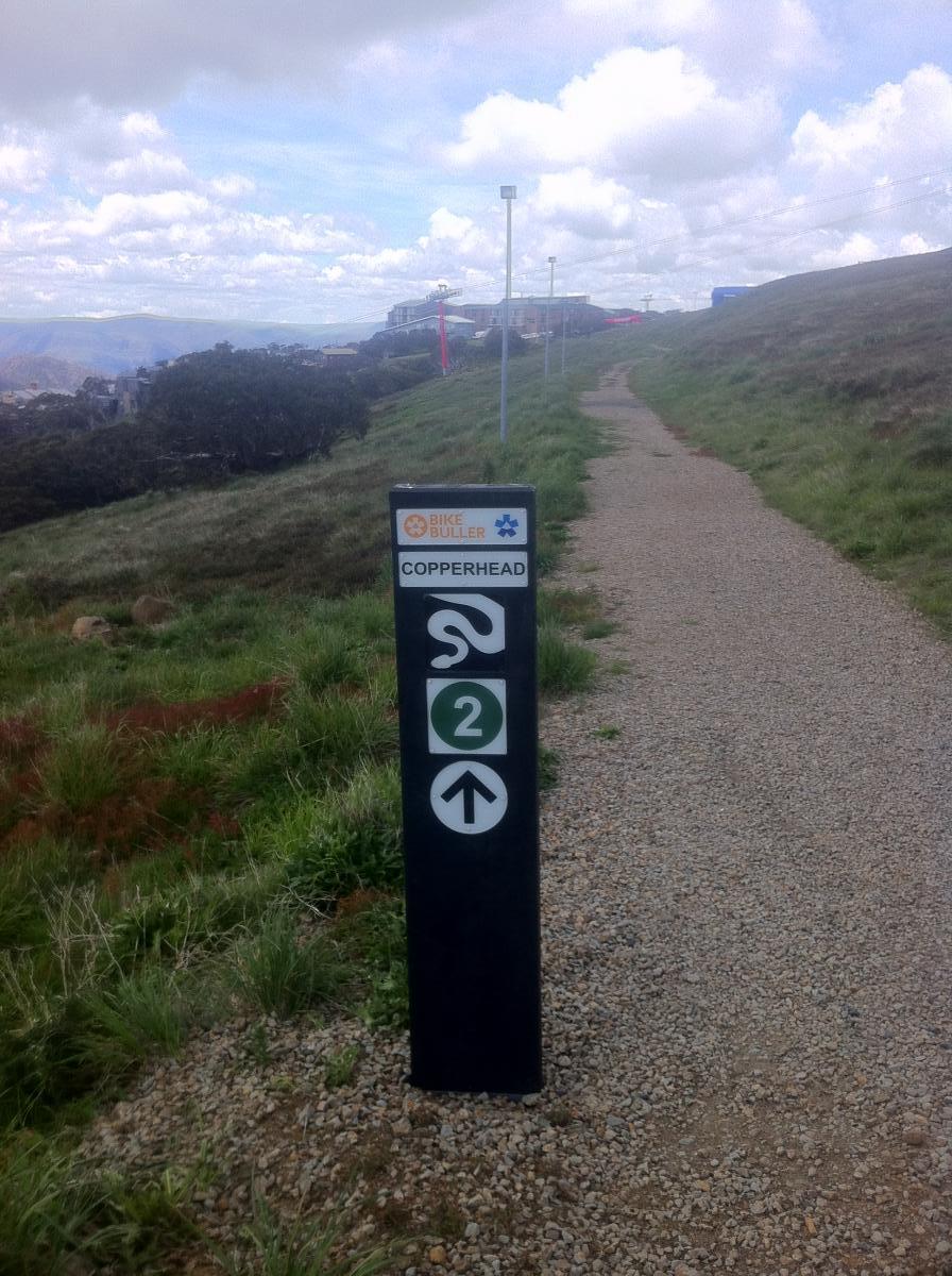 A directional sign for the Copperhead bike trail, featuring a winding path symbol, a "2" for difficulty rating, and an upward arrow. The sign is located on a gravel path surrounded by green grass and hills under a cloudy sky. In the background, a ski resort can be seen on the hillside. Copperhead mountain bike trail.
