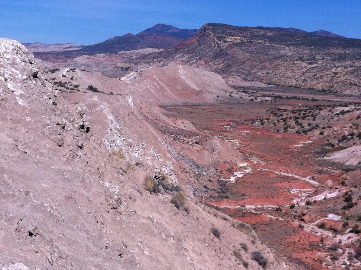A panoramic view of a rugged landscape featuring reddish-brown hills and rocky formations under a clear blue sky. The terrain is marked by dry vegetation and winding paths, with distant mountains visible in the background. White Ridge Bike Trails mountain bike trail.