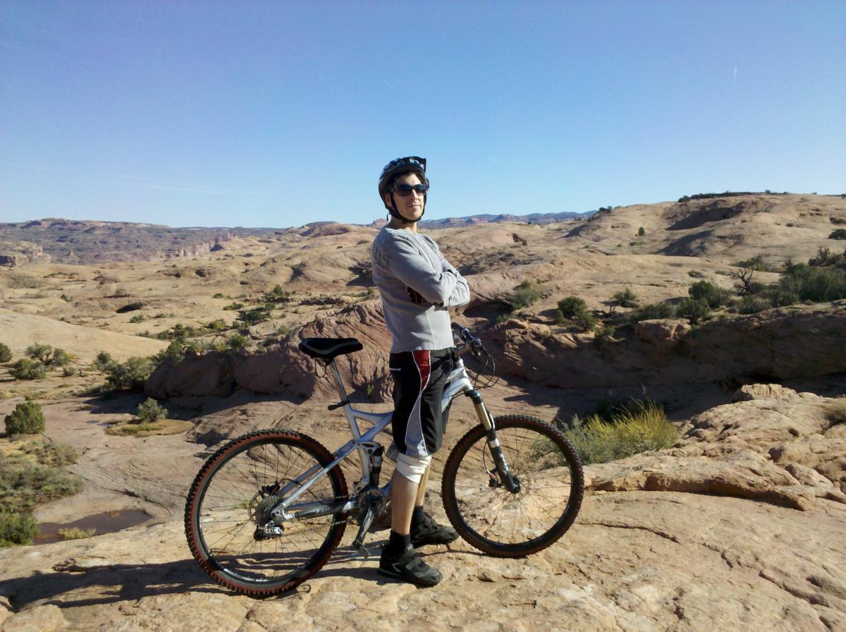 A person wearing a helmet and sunglasses stands confidently next to a mountain bike on a rocky terrain, with expansive desert landscapes in the background under a clear blue sky. Slickrock mountain bike trail.