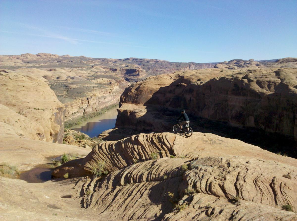 A person riding a mountain bike along a rocky ridge overlooking a winding river and expansive desert landscape under a clear blue sky. Slickrock mountain bike trail.