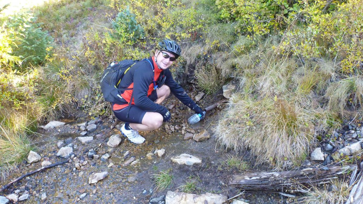 A person in cycling gear crouches beside a stream, reaching down with one hand while holding a water bottle in the other. The surrounding area features tall grass and small bushes, set against a background of autumn foliage. Monarch Crest Trail mountain bike trail.