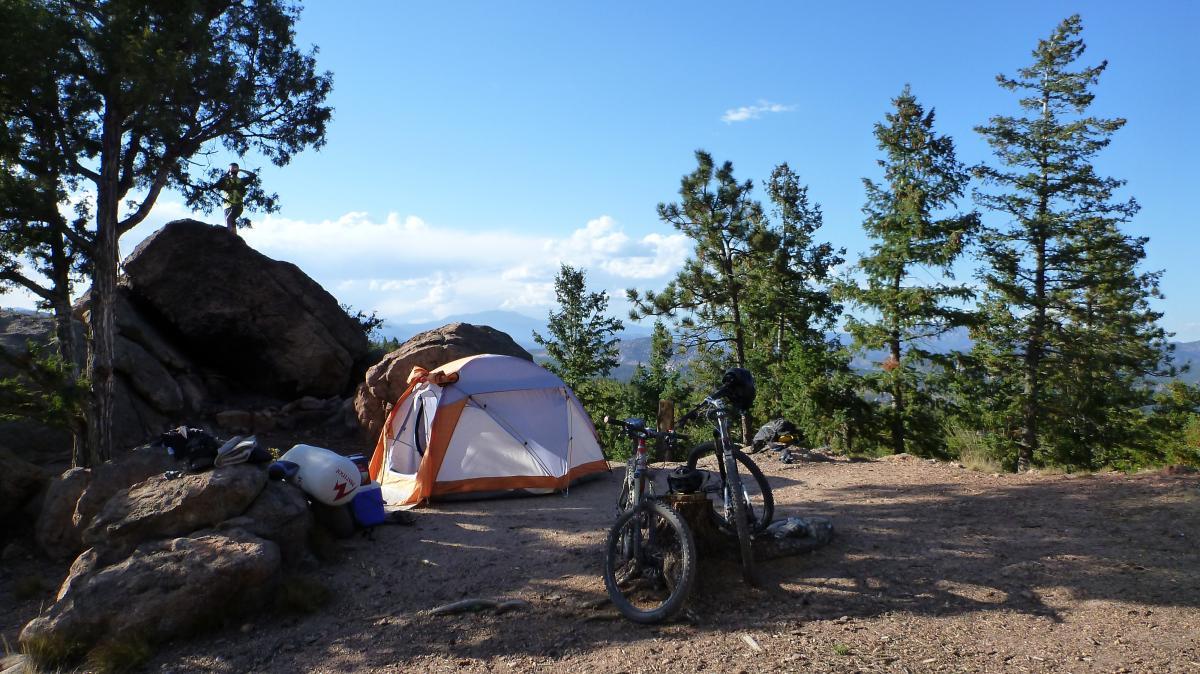 A scenic camping site featuring a tent with orange accents set among rocks and tall pine trees. A mountain bike rests nearby on the ground, and a person is seen climbing on a large boulder in the background. The sky is bright with scattered clouds, creating a serene outdoor atmosphere. Buffalo Creek mountain bike trail.