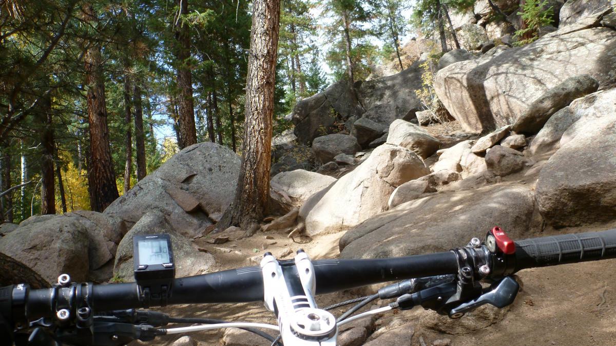 View from the handlebars of a mountain bike on a rocky trail surrounded by trees. The terrain is rough, with large boulders and a dirt path visible. A bike computer is displayed on the handlebars, indicating the rider's stats. Buffalo Creek mountain bike trail.