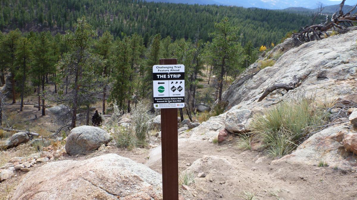 A wooden trail sign marked "THE STRIP" indicating a challenging trail feature ahead, accompanied by iconography illustrating varying levels of difficulty. In the background, a scenic landscape of trees, rocks, and distant mountains under a partly cloudy sky. Buffalo Creek mountain bike trail.