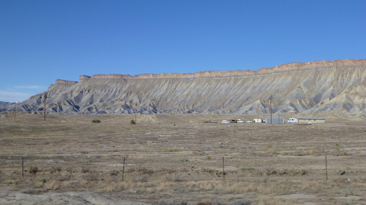 A panoramic view of a rugged, textured landscape featuring a sloping cliff with layered geological formations under a clear blue sky. In the foreground, there are a few buildings and vehicles near the base of the cliff, surrounded by sparse vegetation and open terrain. 18 Road Trails / North Fruita Desert mountain bike trail.