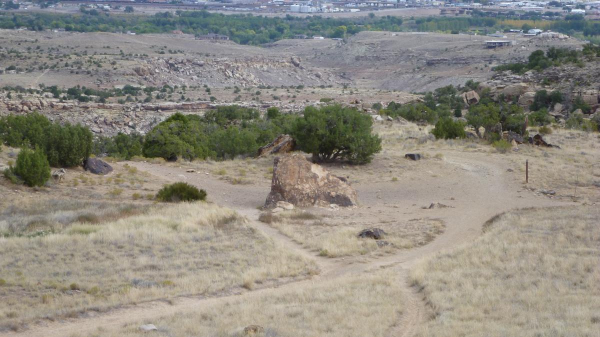 A scenic view of a dry, rocky landscape featuring sparse vegetation, including a few scattered bushes and grassy areas. A prominent rock is situated in the foreground, with a trail leading off to the right, surrounded by hilly terrain and distant structures in the background. Lunch Loops mountain bike trail.