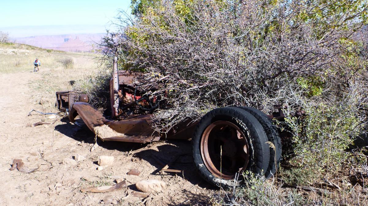 An abandoned, rusted car chassis overgrown with bushes and vegetation in a dry, natural landscape. In the background, a person is seen walking along a dirt path. Porcupine Rim mountain bike trail.