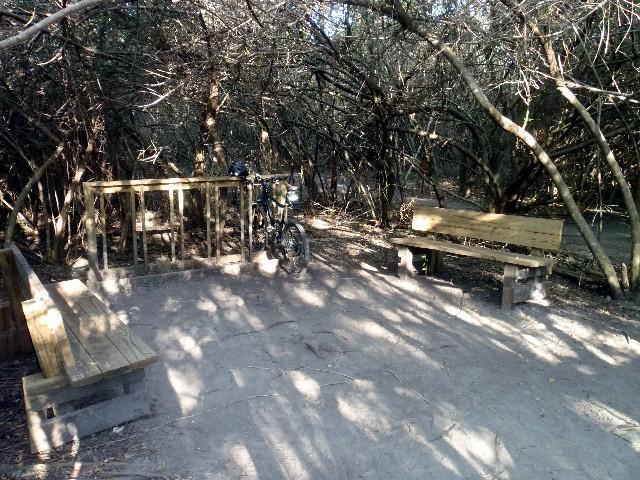 A small, secluded area in a forested setting, featuring two wooden benches and a bike leaning against a railing. The ground is dirt, and the scene is partially shaded by dense foliage overhead, creating a peaceful spot for rest or relaxation amidst nature. Caloosahatchee Regional Park mountain bike trail.