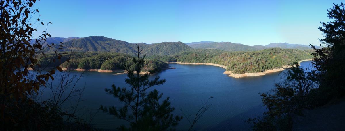 A panoramic view of a tranquil lake surrounded by lush green hills and distant mountains under a clear blue sky. The shoreline is partially visible, showcasing a mix of sandy and rocky areas, while trees frame the scene in the foreground, adding a touch of natural beauty. Tsali Left Loop mountain bike trail.