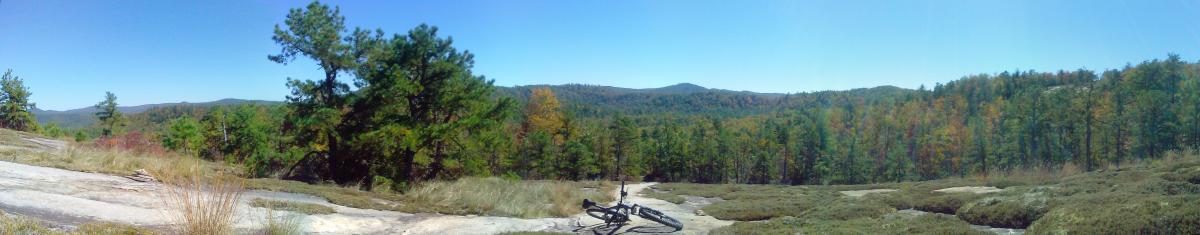 A panoramic view of a scenic landscape featuring rolling hills and a diverse forest in autumn colors. In the foreground, there is an area of rocky terrain and grasses, with a bicycle resting on the ground. The clear blue sky above contrasts with the vibrant green and orange foliage of the trees in the background. Cedar Rock Trail #16 mountain bike trail.