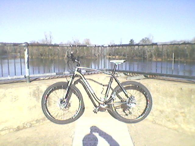 A mountain bike is positioned on a concrete surface near a railing, overlooking a calm body of water surrounded by trees. The scene is set against a clear blue sky. The bike features thick tires and a sleek frame, indicating it is designed for off-road riding. Augusta Canal mountain bike trail.