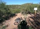 A mountain bike lying on a dirt trail surrounded by desert vegetation, with a signpost in the background. The scene is set under a clear blue sky, showcasing a natural landscape ideal for outdoor activities. Robles Pass mountain bike trail.