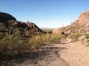 A desert landscape featuring a narrow dirt path flanked by rocky hills. The scene is bathed in bright sunlight, with sparse vegetation and clear blue skies in the background. Robles Pass mountain bike trail.