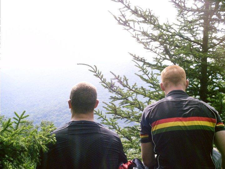 Two individuals sit on a ledge overlooking a vast mountainous landscape, surrounded by evergreen trees. The sunlight casts a bright glow in the background, creating a serene atmosphere. One person is wearing a black shirt, while the other has a shirt featuring horizontal stripes in red, yellow, and green. Gauley Headwaters mountain bike trail.