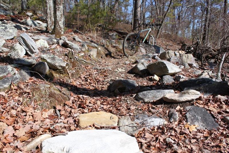A rocky trail through a wooded area, covered in fallen leaves, with a mountain bike partially visible in the background. The scene depicts a natural landscape, suggesting a hiking or biking path. Cheaha State Park mountain bike trail.