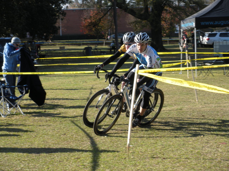 Two cyclists navigate a tight turn on a grassy track marked with yellow tape during a race. The athletes are focused and competitive, wearing helmets and jerseys. In the background, there are spectators and parked vehicles, suggesting a lively outdoor event.