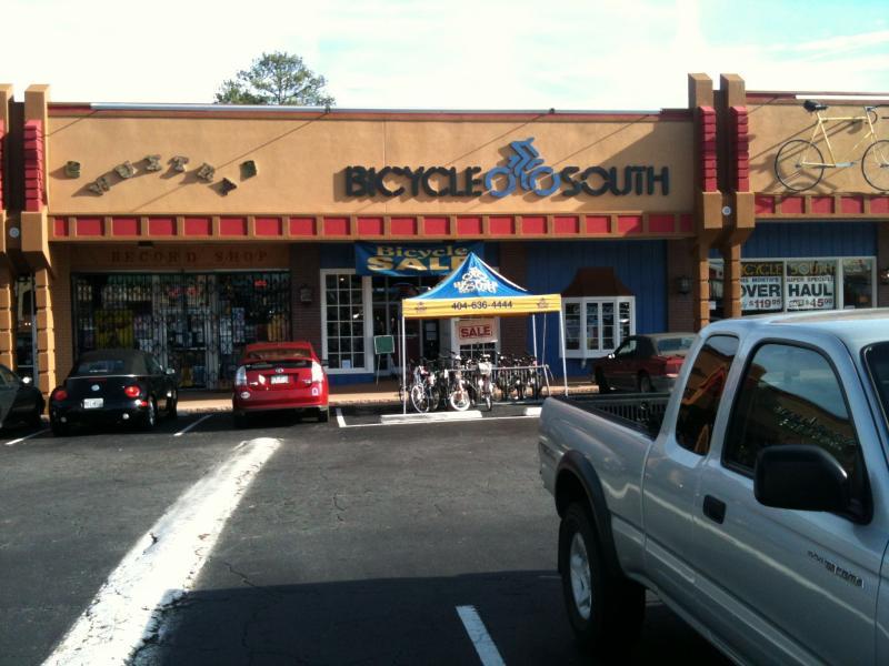 Front view of a store called "Bicycle South," featuring a yellow tent advertising a bicycle sale. The storefront is colorful, with large signs and a decorative bicycle graphic. Several bicycles are displayed outside on the pavement in front of the store, alongside parked cars. A record shop is visible next to the bicycle shop.