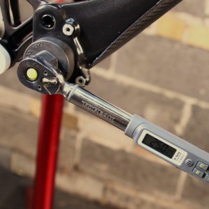 A close-up view of a digital torque wrench attached to a bicycle component, set against a textured wall. The wrench displays a torque reading on its screen, indicating precise measurement for bike maintenance.