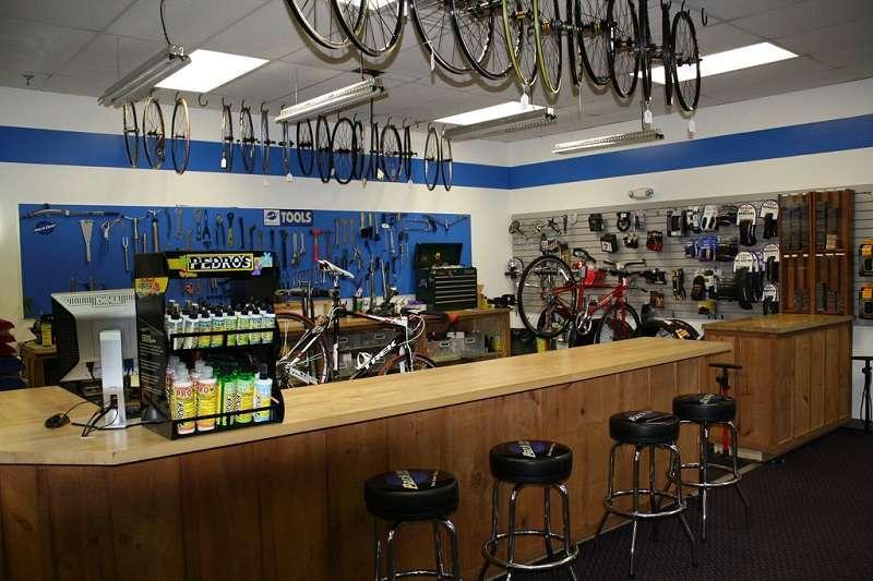 A bicycle shop interior featuring a wooden counter with bar stools. The walls are adorned with hanging bikes and an array of tools on a blue and white background. Various biking accessories and products are displayed on shelves. A cash register and a computer can also be seen on the counter.