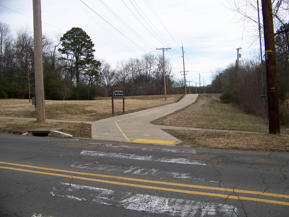 A view of a paved pathway leading from a road to a grassy area, with sparse trees in the background and utility poles lining the scene. A sign labeled "Recreational Trail" is visible near the start of the path. The sky is cloudy, and the landscape appears to be in a suburban setting. Spadra Creek Nature Trail mountain bike trail.