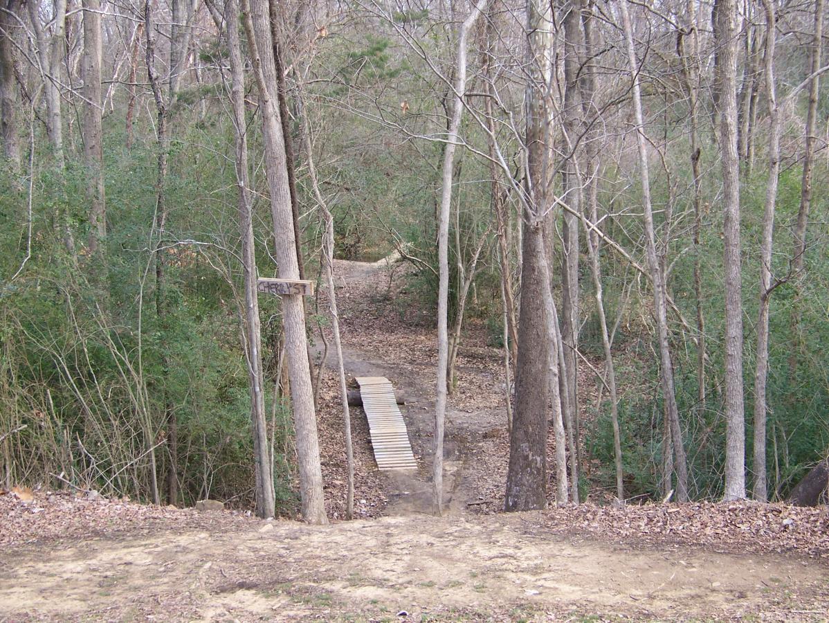 A wooded trail with a wooden plank bridge crossing a small path. The scene features bare trees and scattered leaves on the ground, suggesting a serene, natural setting. A sign labeled "CHRIST" is visible on a tree near the bridge. Spadra Creek Nature Trail mountain bike trail.