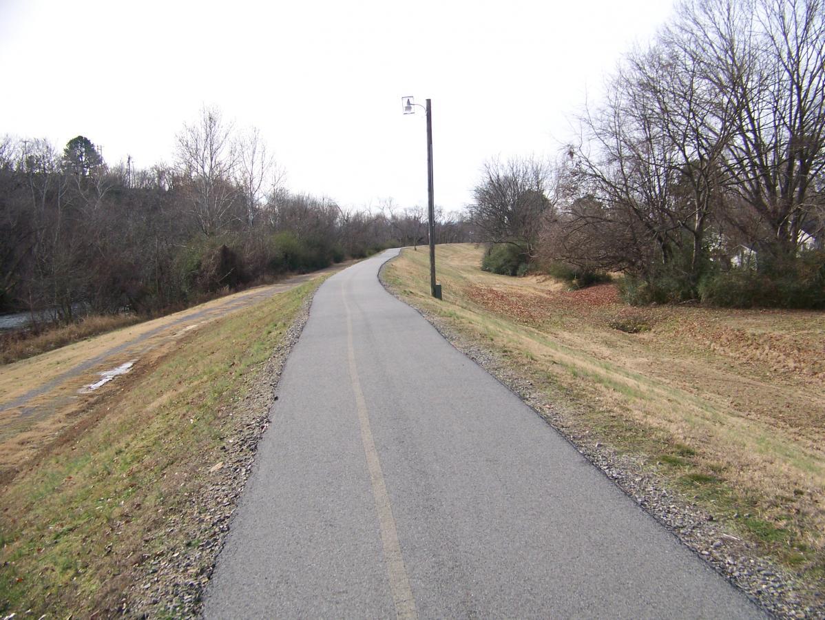 A winding paved path runs alongside a grassy slope, bordered by bare trees on either side. A telephone pole stands next to the path, while a small stream can be seen on the left side. The scene is set against a cloudy sky, suggesting a cool day in a natural setting. Spadra Creek Nature Trail mountain bike trail.