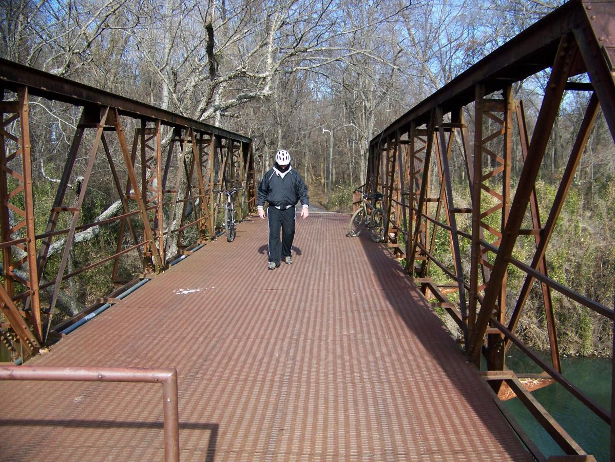 A person wearing a helmet and a black jacket walks across a metal bridge surrounded by bare trees. A bicycle is parked on the side of the bridge. The scene is set in a natural environment, highlighting the bridge's rusted structure and the serene surroundings. Spadra Creek Nature Trail mountain bike trail.