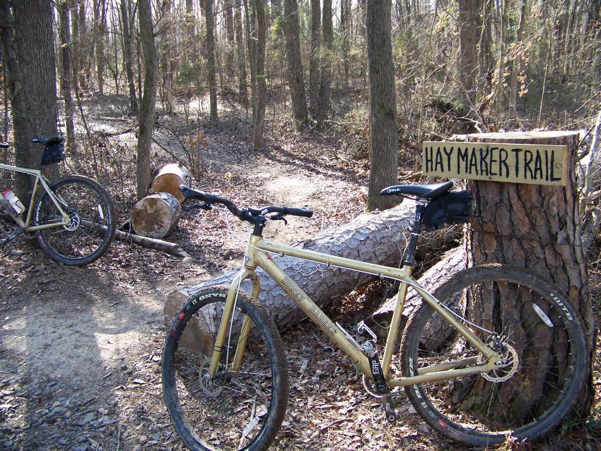 Two mountain bikes are parked on the side of a dirt trail in a forest. A sign labeled "HAY MAKER TRAIL" is attached to a tree nearby. The area is surrounded by leafless trees, suggesting it is early spring or late fall, with scattered leaves on the ground. A fallen log lies across the trail, enhancing the natural, rustic setting. Spadra Creek Nature Trail mountain bike trail.