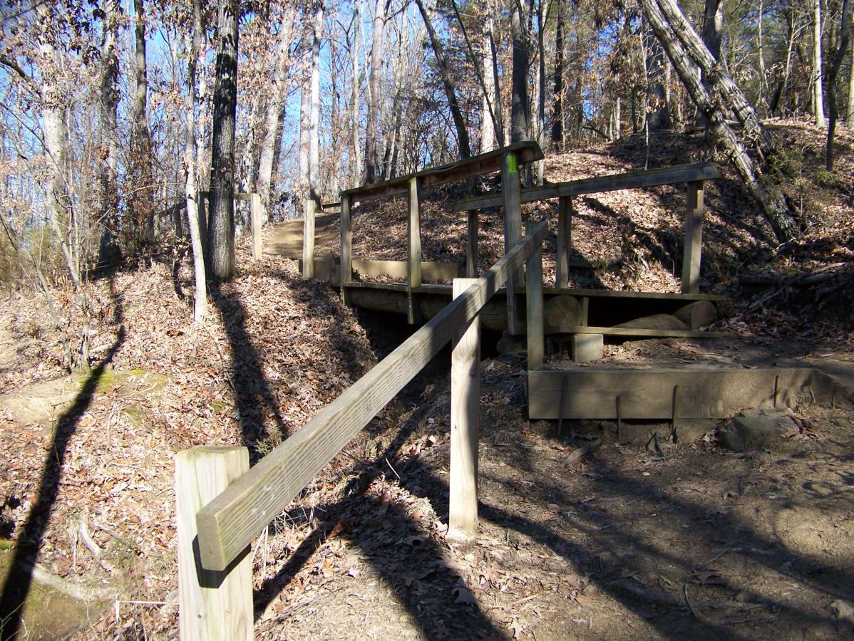 A wooden bridge crosses a small incline in a forested area, surrounded by trees and scattered dry leaves on the ground. The scene is illuminated by sunlight filtering through the branches, creating a serene outdoor atmosphere. Spadra Creek Nature Trail mountain bike trail.