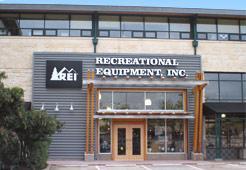 The image shows the exterior of an REI (Recreational Equipment, Inc.) store, featuring a large sign with the company name. The building has a modern design with wooden accents and large windows.