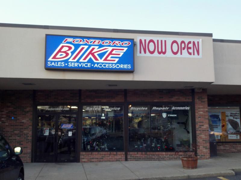 Image of a bicycle shop with a sign that reads "Foxboro BIKE" and "NOW OPEN." The storefront features glass doors showcasing bicycles inside, along with a sign highlighting "Sales, Service, Accessories." The building has a brick facade and is located in a plaza.