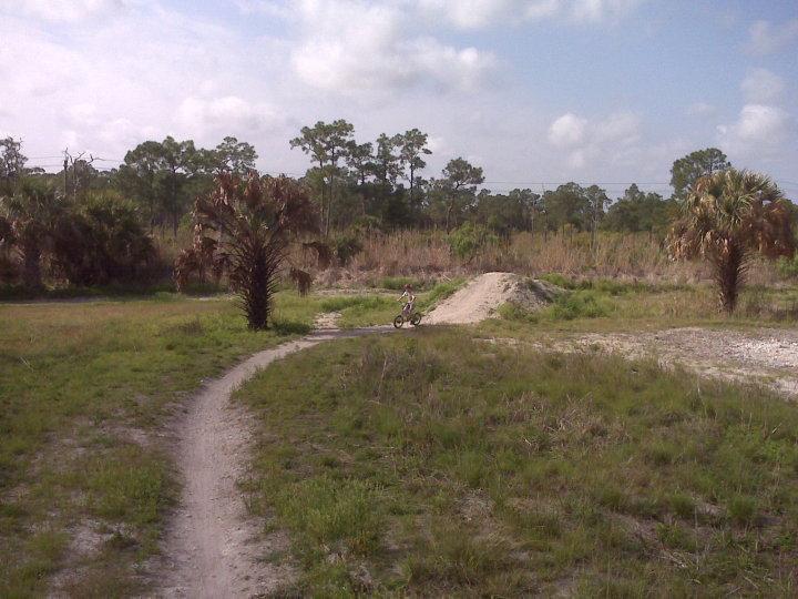 A dirt path winds through a grassy area dotted with palm trees, leading to a small dirt ramp in the background. A child can be seen riding a bicycle along the path, enjoying the outdoors under a partly cloudy sky. Caloosahatchee Regional Park mountain bike trail.
