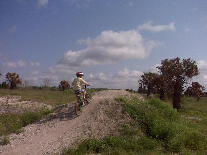 A child riding a bicycle on a dirt hill in an open field, with palm trees in the background and a blue sky with fluffy clouds. Caloosahatchee Regional Park mountain bike trail.