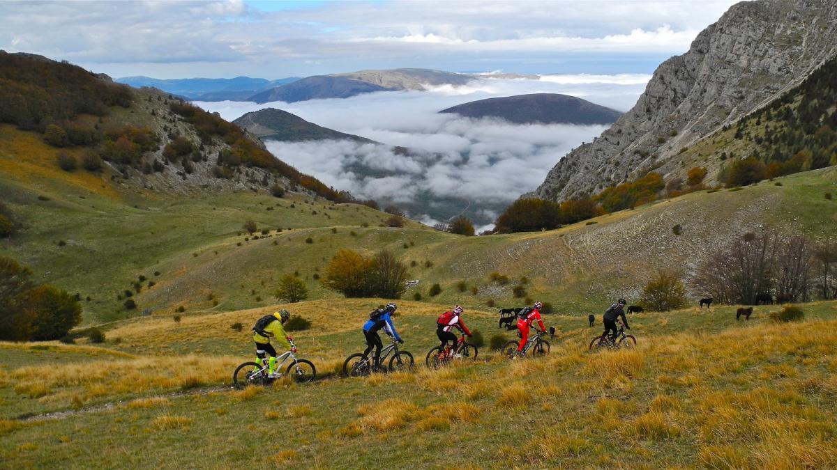 A group of six mountain bikers rides along a grassy trail in a picturesque landscape. The scene features rolling hills, rocky outcrops, and a backdrop of mountains partially shrouded in clouds. Autumn foliage adds splashes of color to the hillsides, with a few cows grazing in the distance. The sky is overcast, creating a moody yet serene atmosphere. Val Di Bove mountain bike trail.