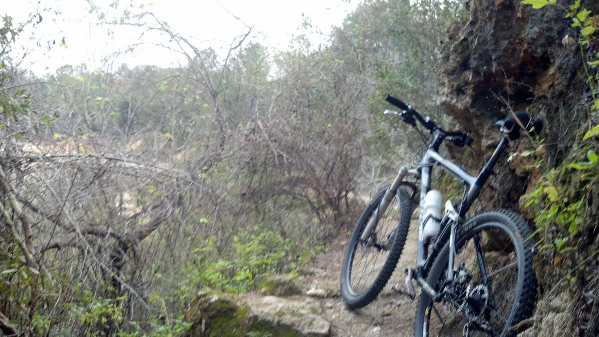 A mountain bike parked on a narrow trail surrounded by dense vegetation and rocky terrain, with a hint of a canyon visible in the background. Imba Red mountain bike trail.