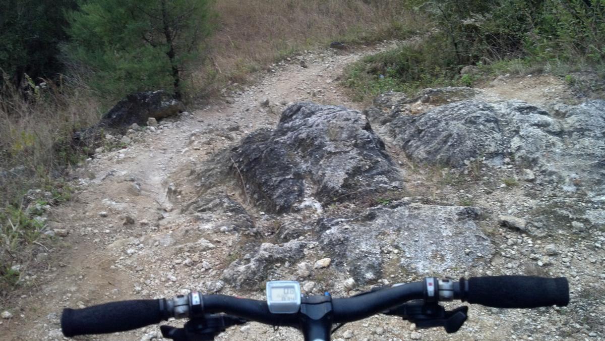 A close-up view from the handlebars of a mountain bike, looking down a rocky trail surrounded by grass and small trees. The path is uneven, with large rocks and loose gravel, indicating a challenging terrain for cycling. A display on the bike's handlebars shows speed or distance metrics. Imba Red mountain bike trail.