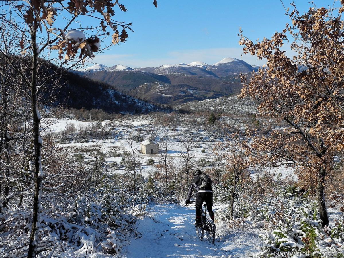 A cyclist riding through a snowy landscape with mountains in the background and a small chapel in the foreground. The scene is framed by trees with some snow on their branches, showcasing a clear blue sky on a sunny day. Macereto mountain bike trail.