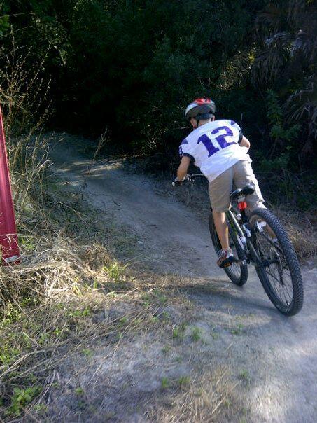 A child wearing a helmet and a white jersey with the number 12 is riding a mountain bike down a dirt trail surrounded by greenery. The trail slopes downward, and the sun illuminates the scene, highlighting the bike's path. Caloosahatchee Regional Park mountain bike trail.