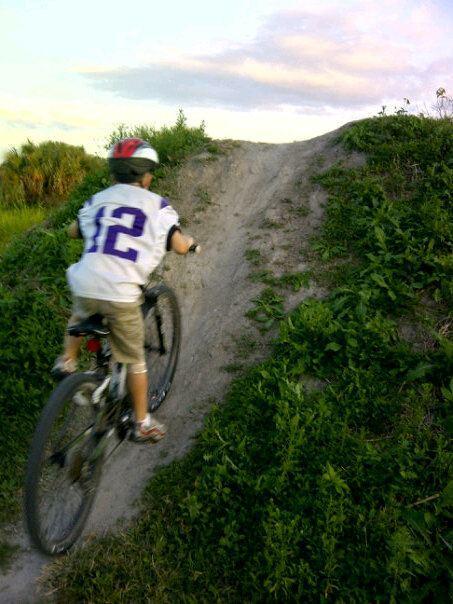 A child riding a bicycle up a dirt ramp surrounded by greenery, wearing a helmet and a sports jersey. The sky is partly cloudy, and the sun is setting in the background. Caloosahatchee Regional Park mountain bike trail.