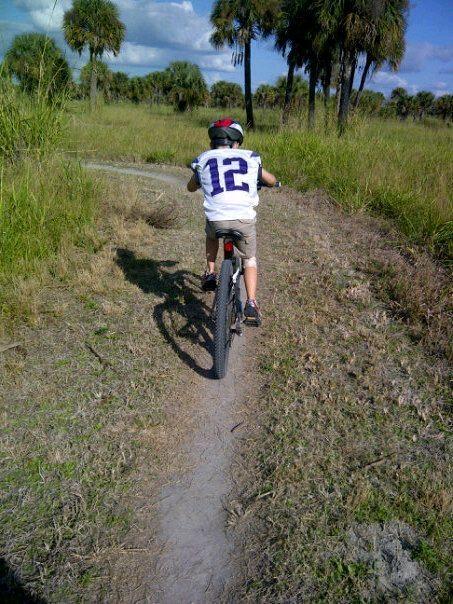 A child riding a bicycle along a dirt path in a grassy area, wearing a helmet and a white shirt with the number 12. The background features tall grass and palm trees under a blue sky with a few clouds. Caloosahatchee Regional Park mountain bike trail.