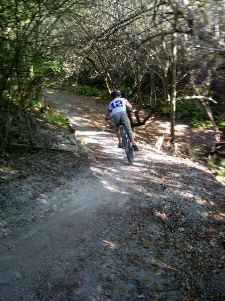 A person riding a mountain bike down a gravel trail surrounded by trees. The rider is wearing a helmet and a shirt with the number 12 on the back. Sunlight filters through the branches, illuminating the path ahead. Caloosahatchee Regional Park mountain bike trail.