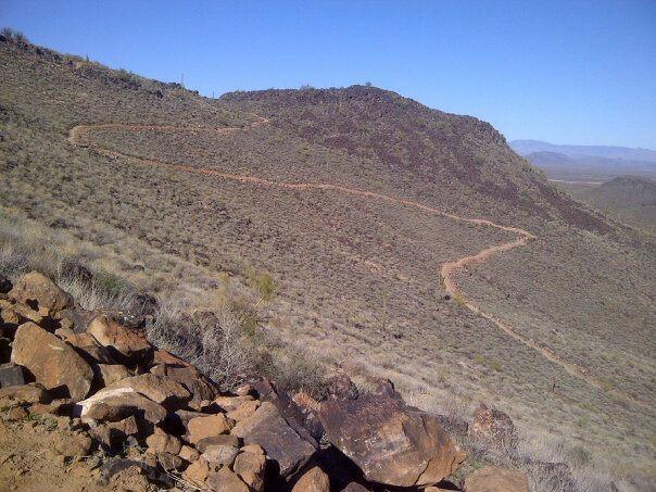 Winding dirt trail leading up a mountainous landscape, with rocky terrain and sparse vegetation under a clear blue sky. Deem Hill mountain bike trail.