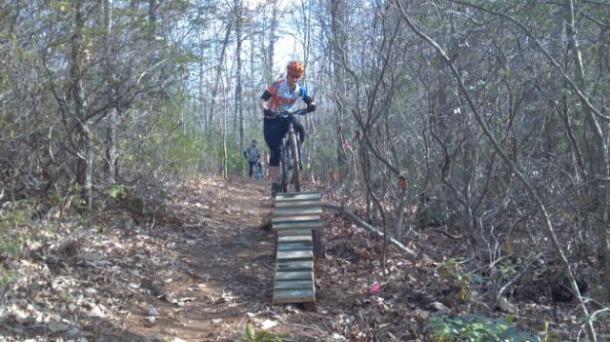 A mountain biker performing a jump off a wooden ramp on a forested trail, surrounded by trees and vegetation, with another cyclist visible in the background. Woolwine Trails [Shiners Revenge] mountain bike trail.