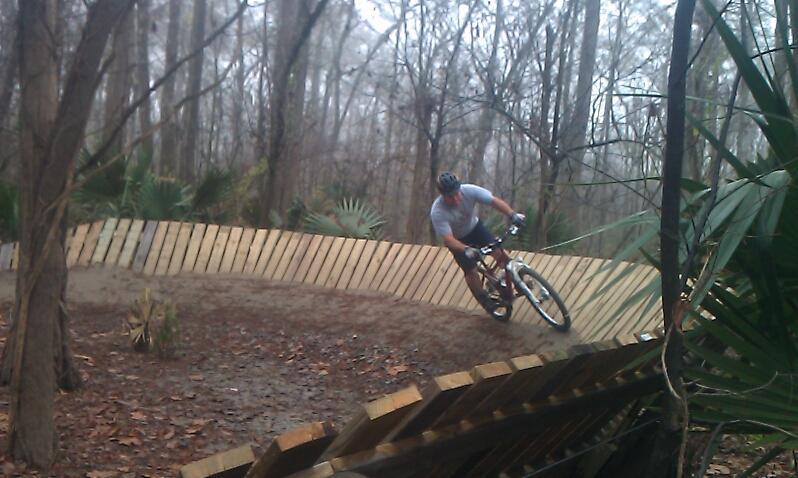 A cyclist leaning into a turn on a wooden track in a wooded area, surrounded by trees and plants. The scene conveys a sense of motion and adventure in a misty environment. Bonnet Carre Spillway Trail mountain bike trail.