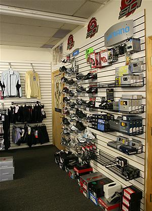 A retail store interior featuring a wall display with various bicycle accessories and gear. The display includes shelves stocked with items such as helmets, lights, and audio equipment, alongside clothing hung on adjacent racks.